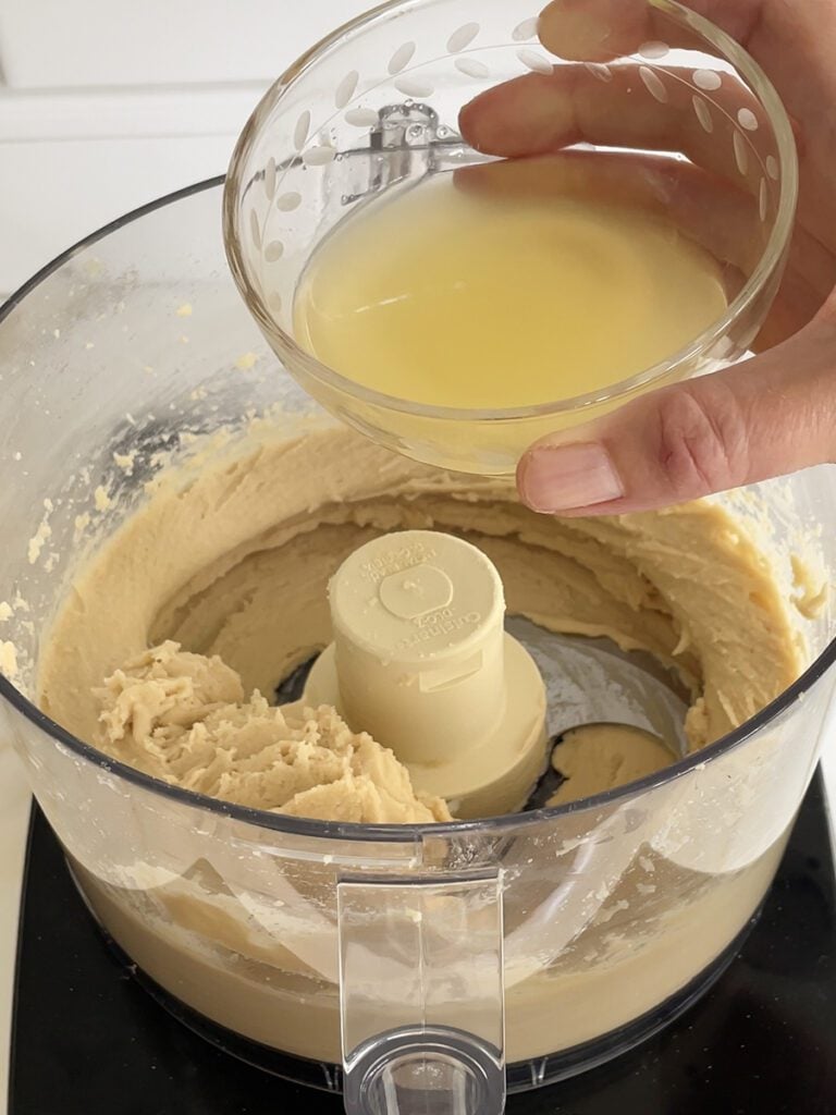 A hand pouring some lemon juice from a glass bowl into the food processor.