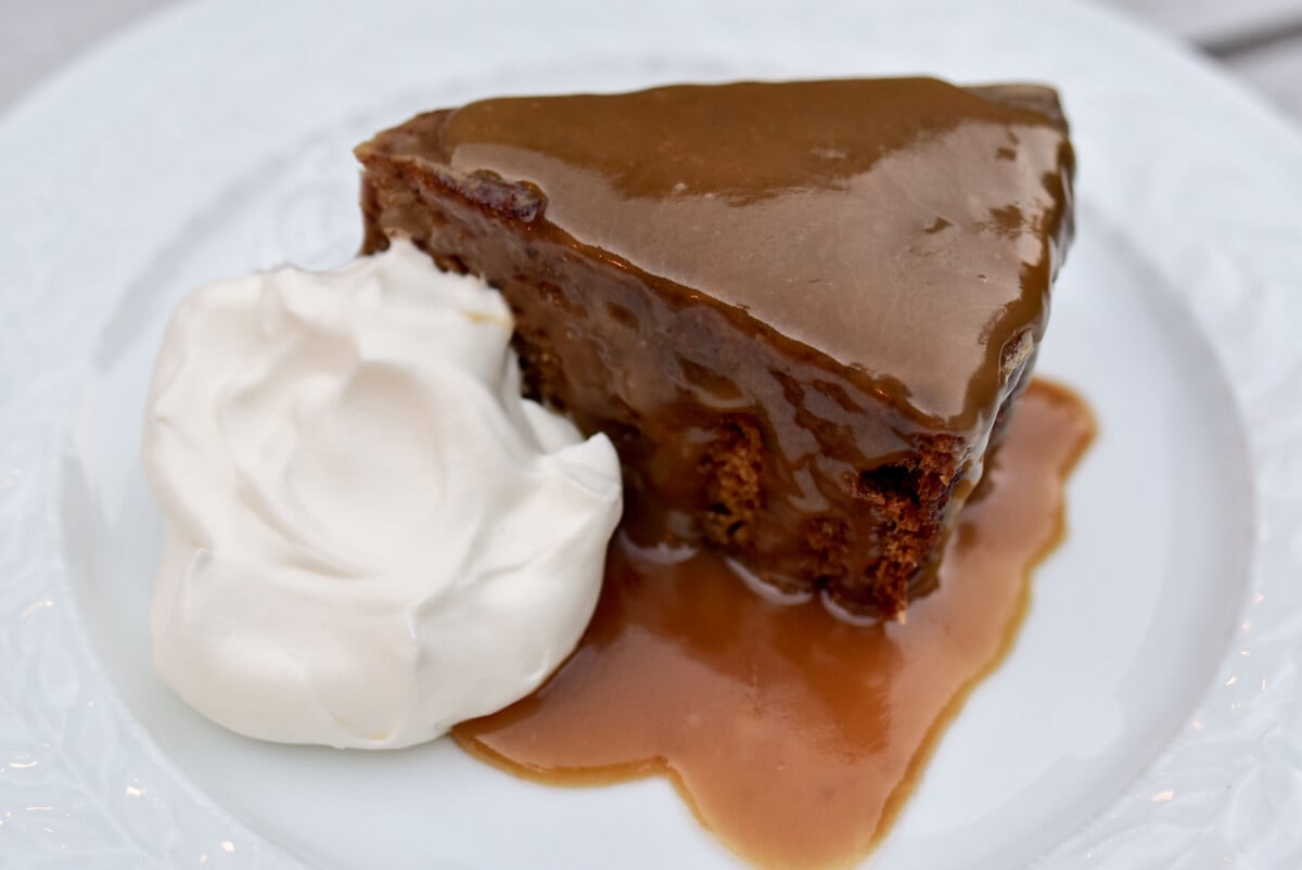 A wedge of brown sticky date cake covered with glossy caramel sauce and a dollop of whipped cream