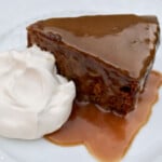 A wedge of brown sticky date cake covered with glossy caramel sauce and a dollop of whipped cream