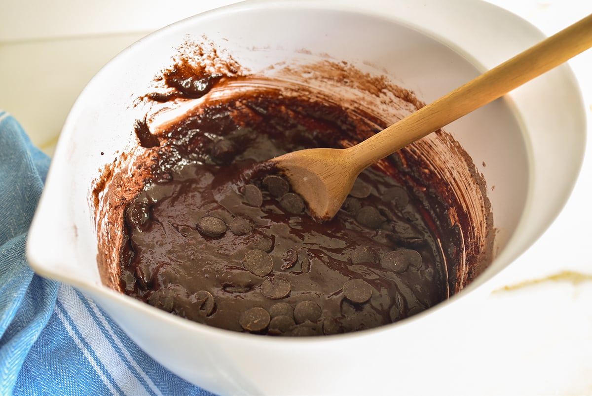 Brownie batter in a white bowl with a wooden spoon and blue towel