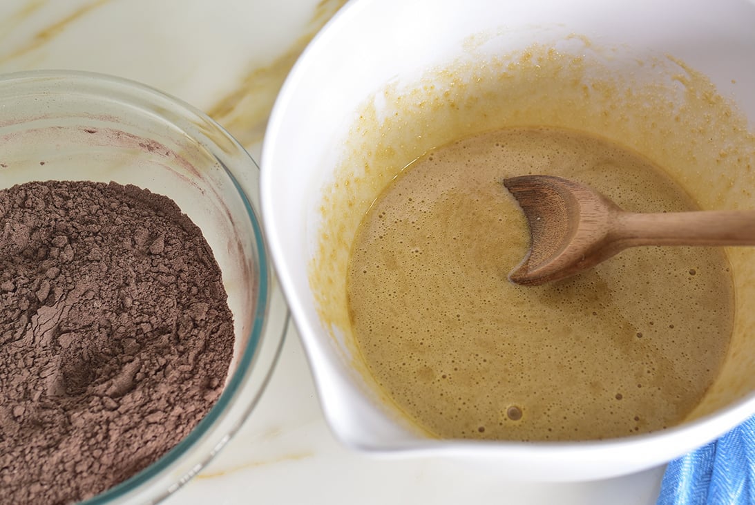 Two bowls side by side, one with creamy batter and a wooden spoon and the other with chocolate colored dry ingredients