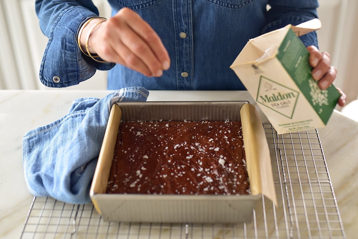 A hand sprinkling flakey sea salt over baked brownies in the pan