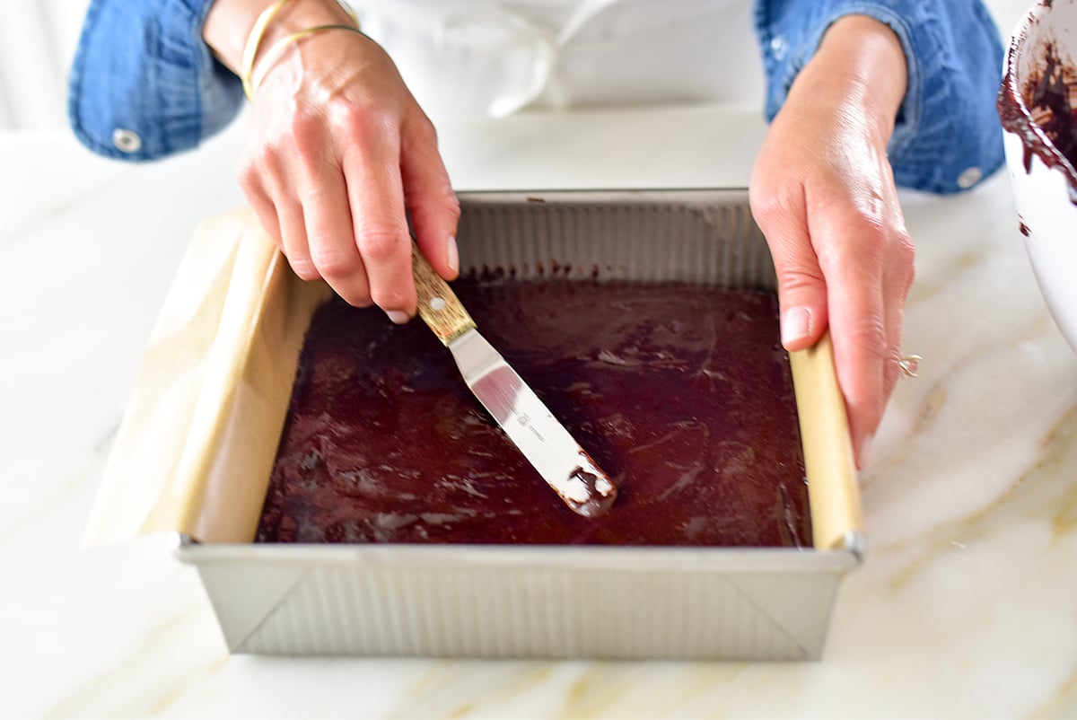 Two hands spreading brownie batter with an offset spatula