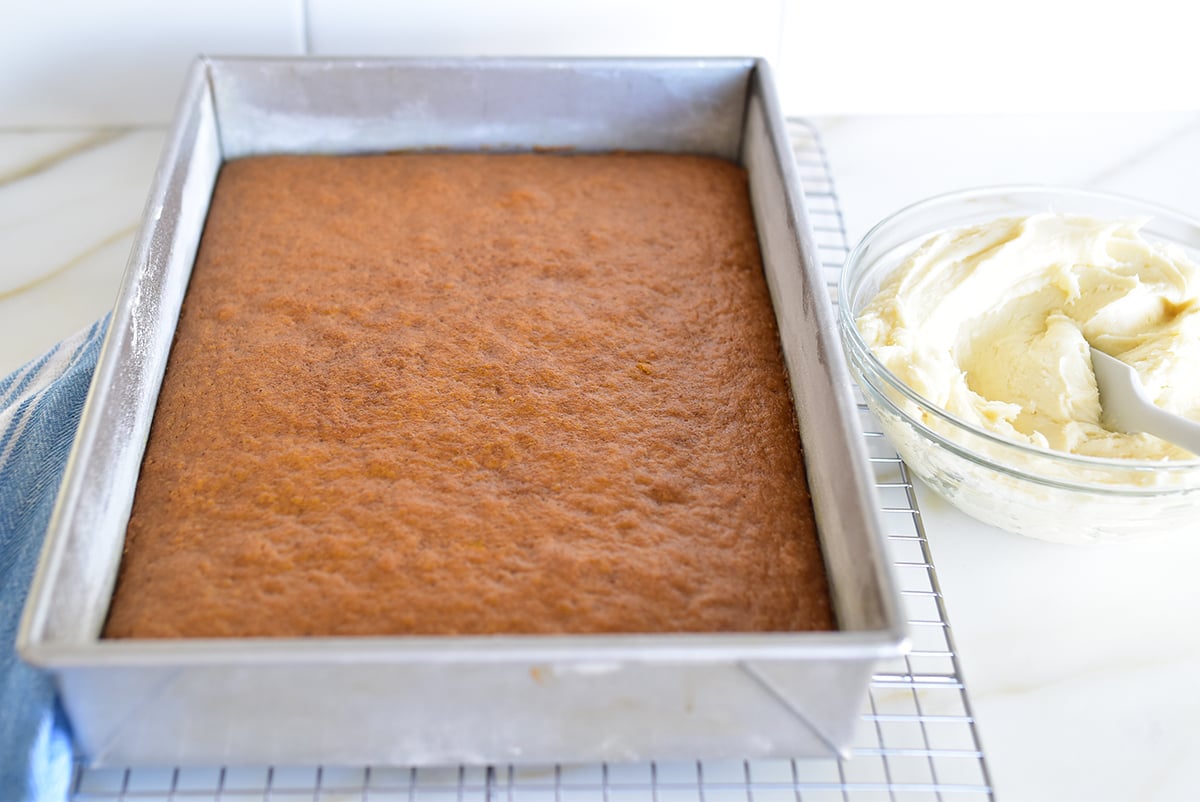 Baked pumpkin bars in a metal pan with white frosting in a glass bowl beside it