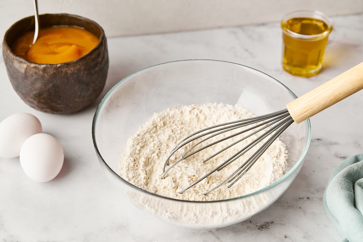 A bowl of flour with a whisk in it and other ingredients around it