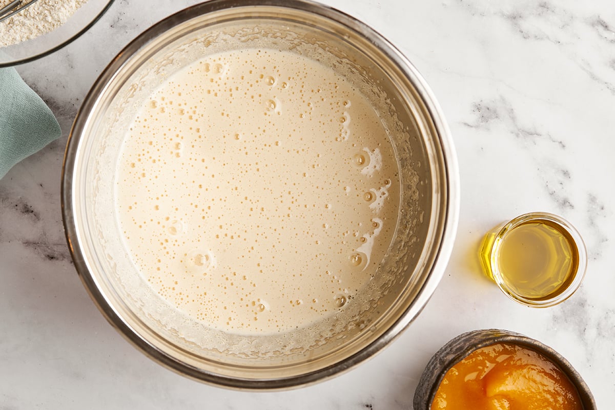 A bowl with wet ingredients combined in it with pumpkin and oil on the side on a marble surface