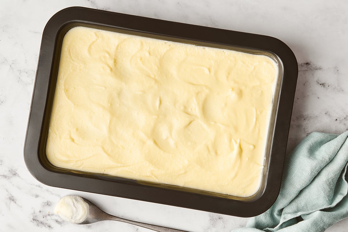 White frosting on a cake in a dark cake pan on a marble surface with light blue towel on the side