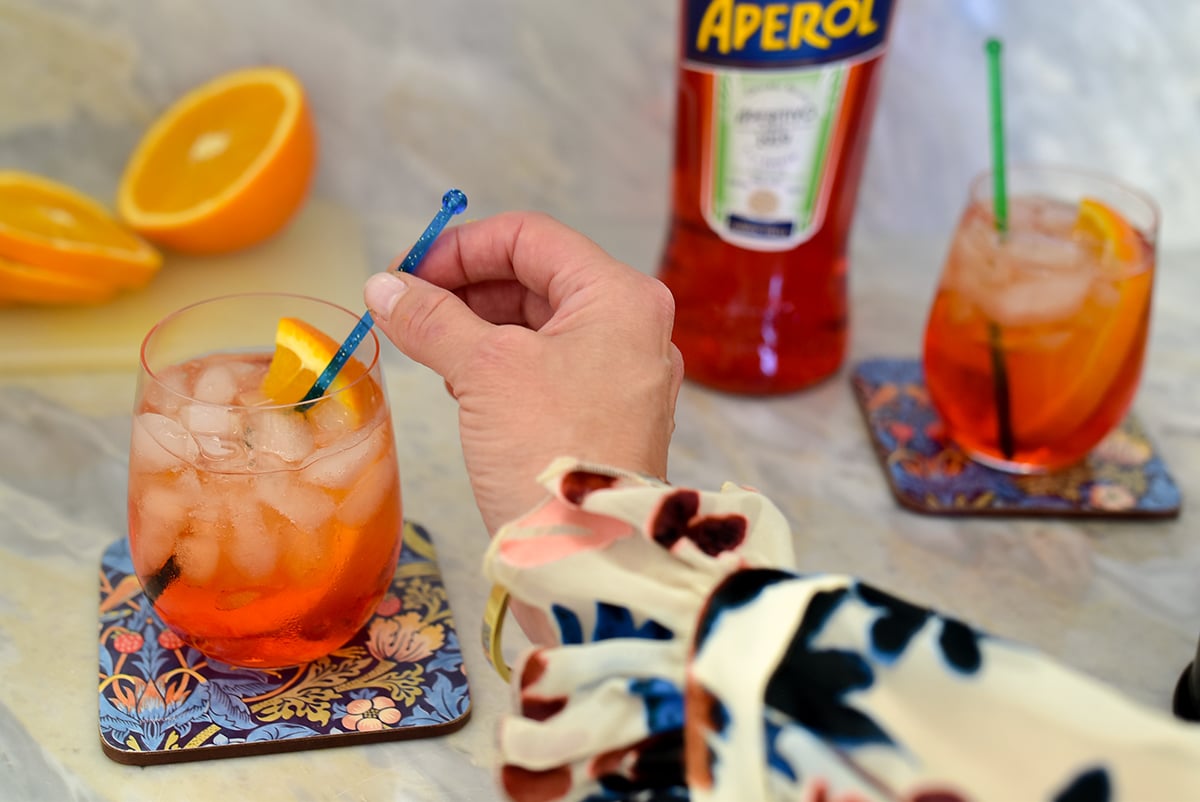 Aperol Spritz cocktail with a hand sitrring it with a blue stirrer