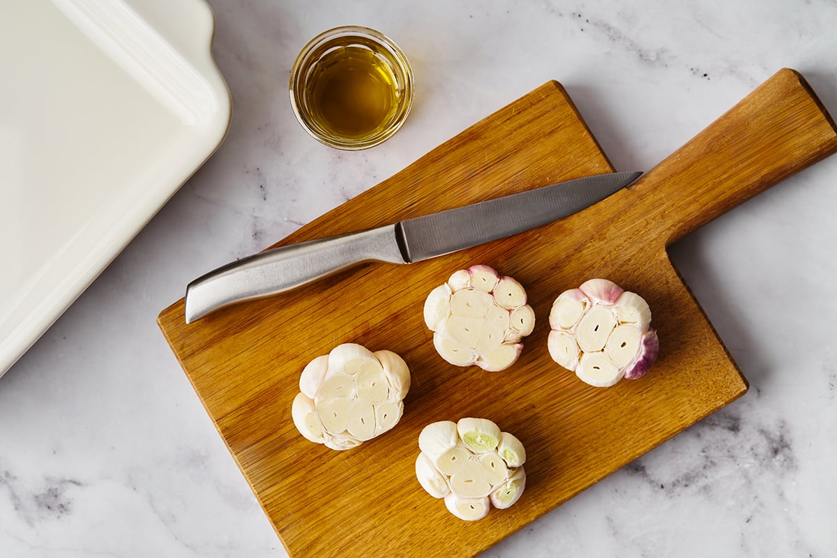 Garlic cloves with the tops cut off next to a knife on a cutting board