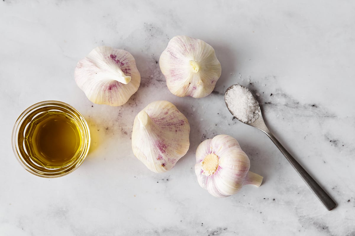 Garlic cloves with a teaspoon of salt and glass bowl of olive oil on a marble surface
