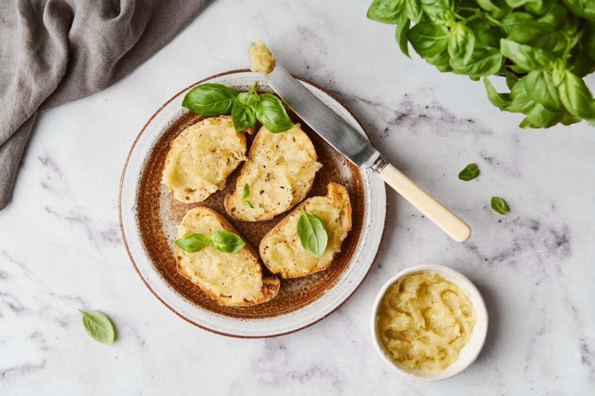 Roasted garlic spread on crostini on a round plate with a knife and basil leaves