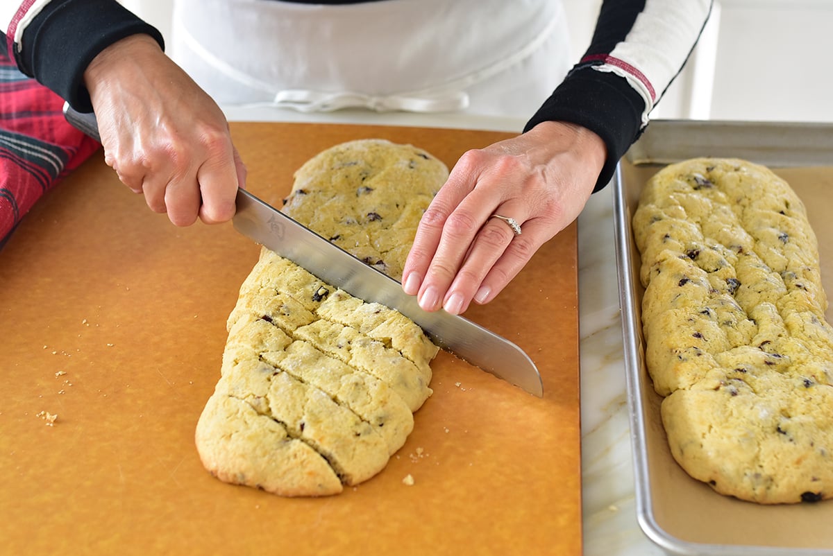 Two hands slicing a biscotti log after the first bake, on the diagonal with a serrated knife