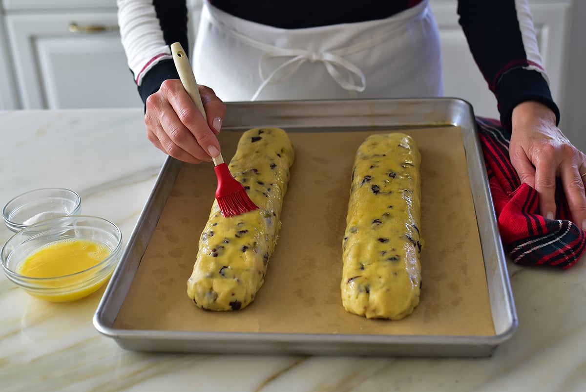 A hand with red silicone brush spreading egg wash on biscotti dough log