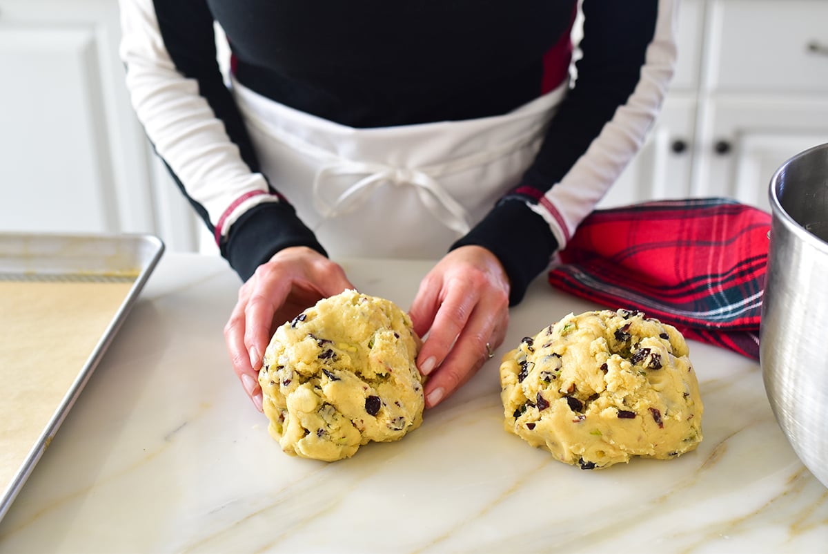 Two hands holding biscotti dough in two balls on the countertop with red towel on the side