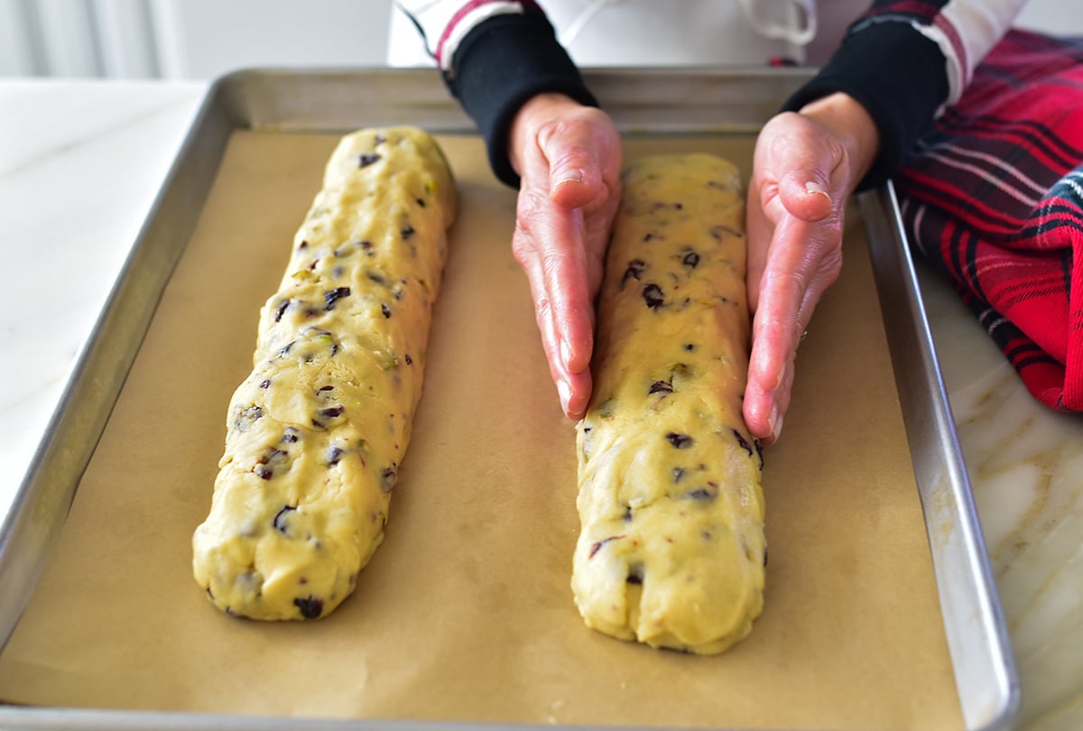 Two hands shaping biscotti logs on a baking sheet