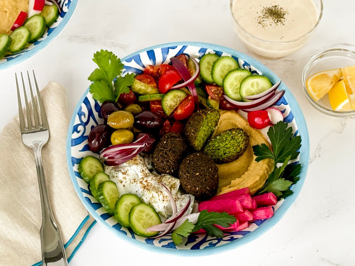 Falafel bowl in a blue and white bowl with a fork beside it along with tahini sauce and lemon slices in small bowls