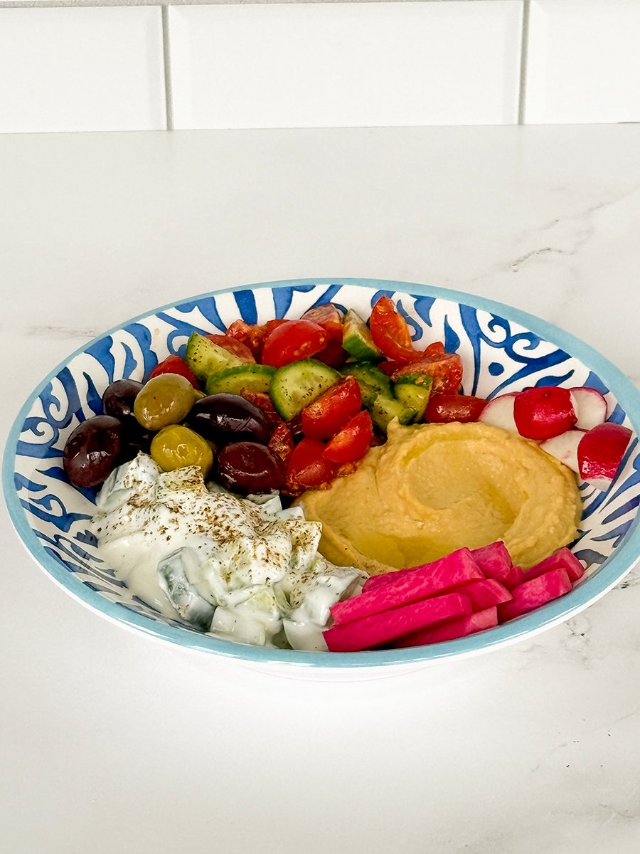 blue and white bowl with hummus, creamy cucumber salad, pink turnips, olives, radishes