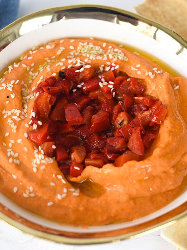 Roasted red pepper hummus in a white bowl with a gold rim surrounded by pita bread