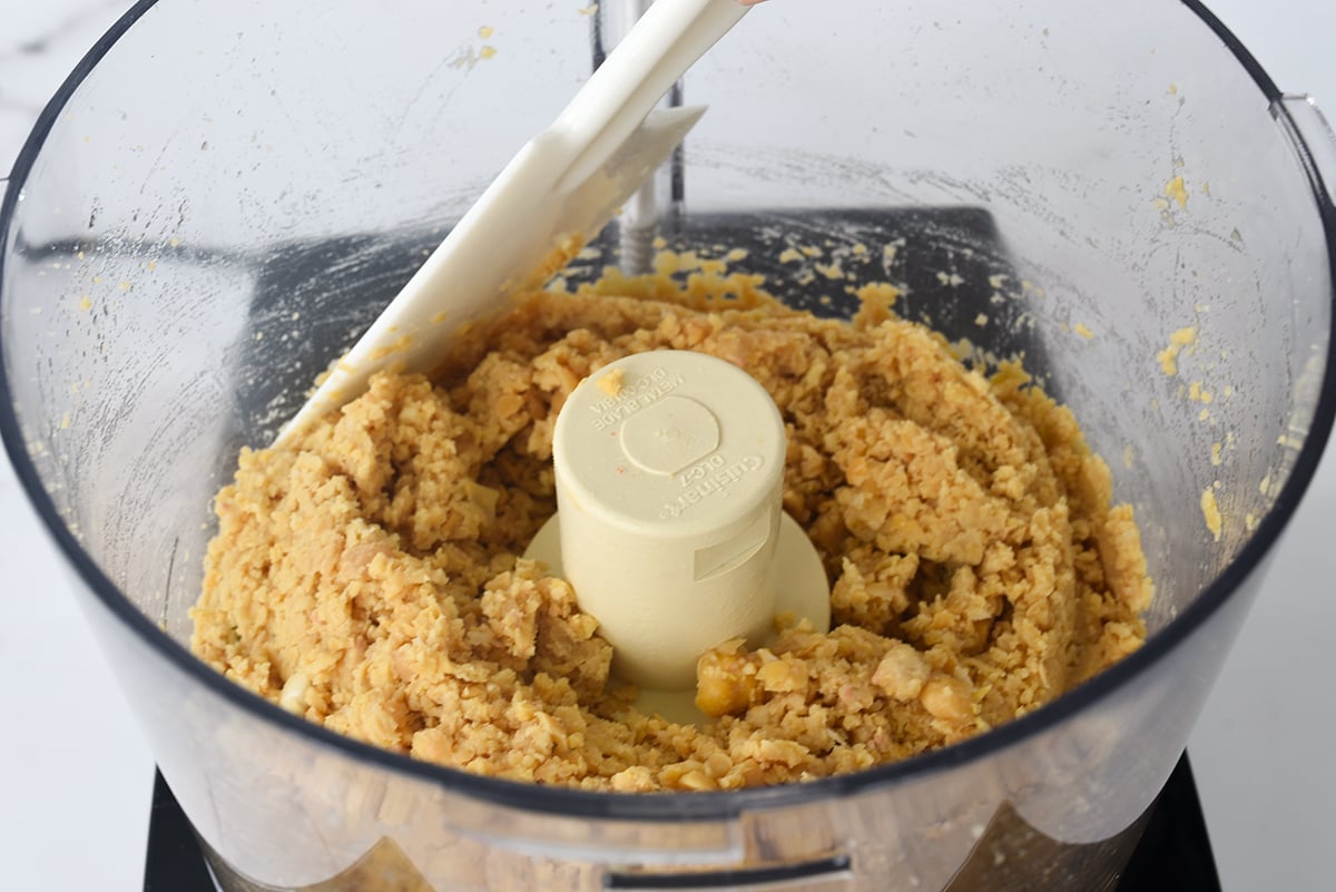 A spatula scraping down the bowl of a food processor with pulverized chickpeas