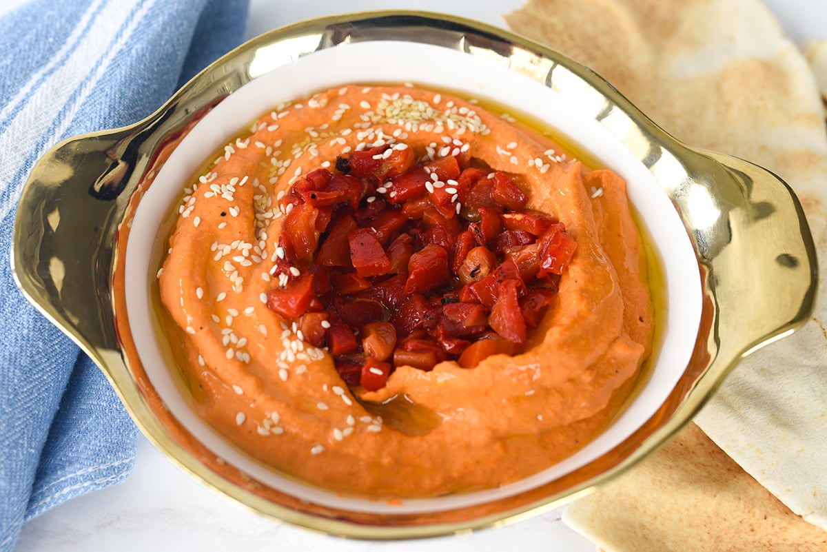 Roasted red pepper hummus in a white bowl with a gold rim surrounded by pita bread