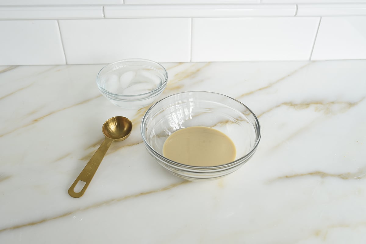 A bowl of tahini with a small bowl of ice water and a gold tablespoon next to it