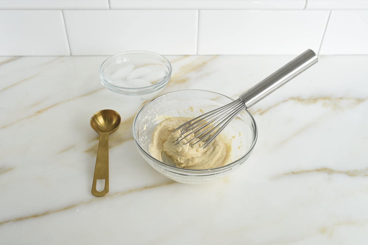 A bowl of tahini with a whisk in it with a small bowl of ice water and a gold tablespoon next to it