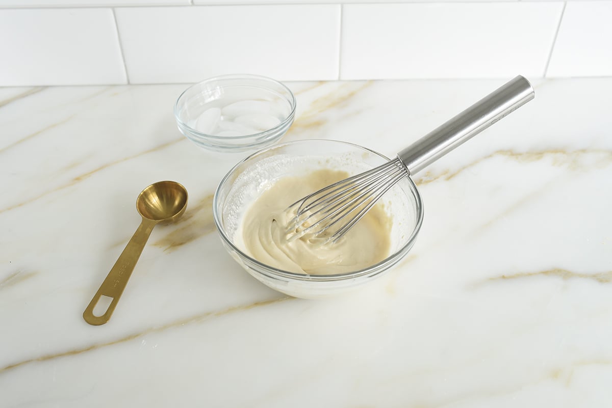 A bowl of smooth tahini with a whisk in it with a small bowl of ice water and a gold tablespoon next to it