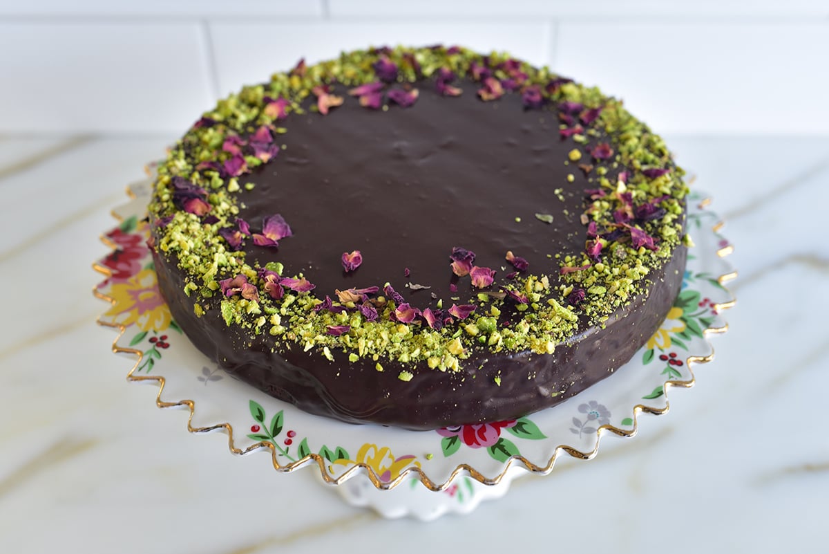 a round chocolate cake on a floral cake plate with pistachios around the edge