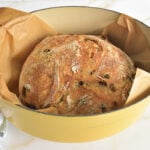 A golden brown loaf of olive bread in a dutch oven lined with parchment