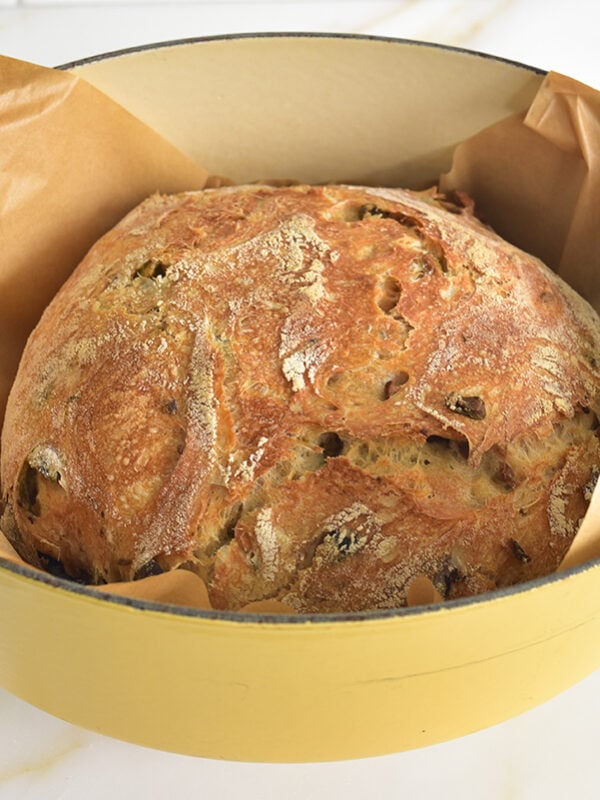 A golden brown loaf of olive bread in a dutch oven lined with parchment