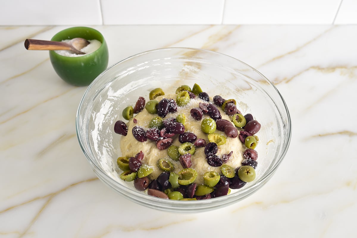 Dough in a glass bowl with chopped olives on top and a green bowl of salt on the side