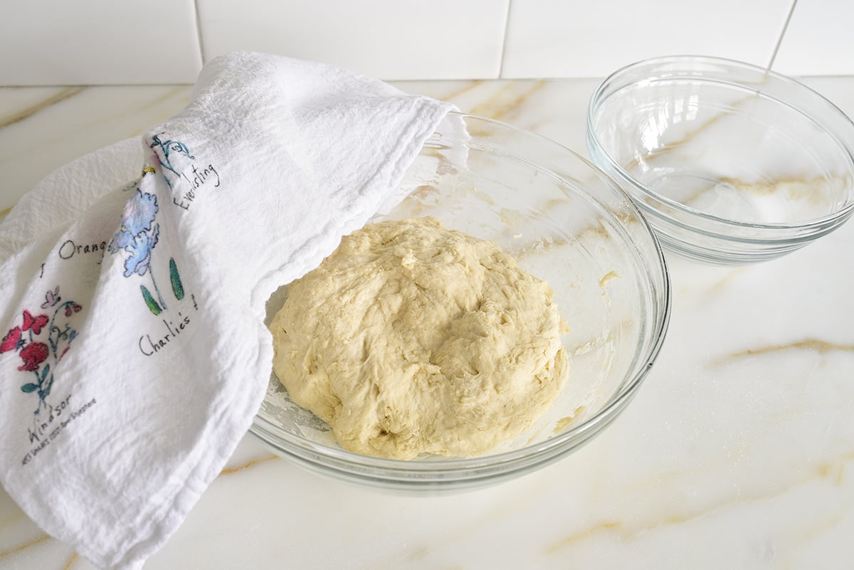 A glass bowl of dough on a marble counter with a towel over the top