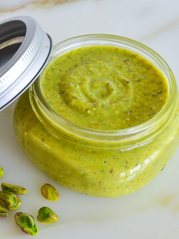 Pistachio cream in a jar with pistachios scattered around it on a marble countertop