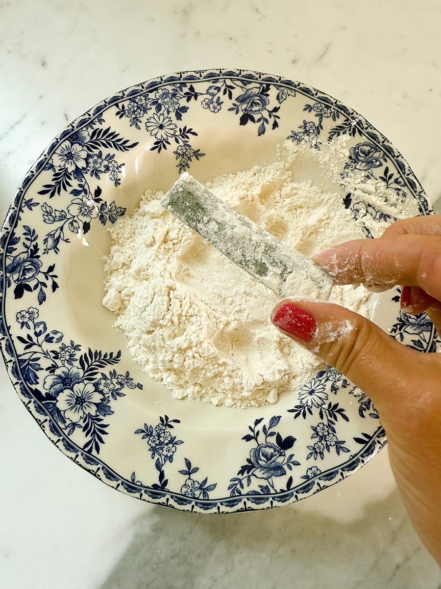Fingers with red nailpolish holding zucchini over a bowl of flour