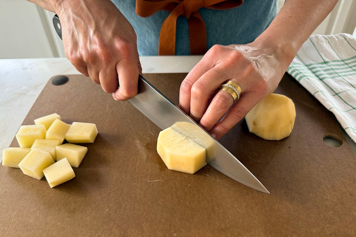 Two hands cubing potatoes with a knife on a cutting board