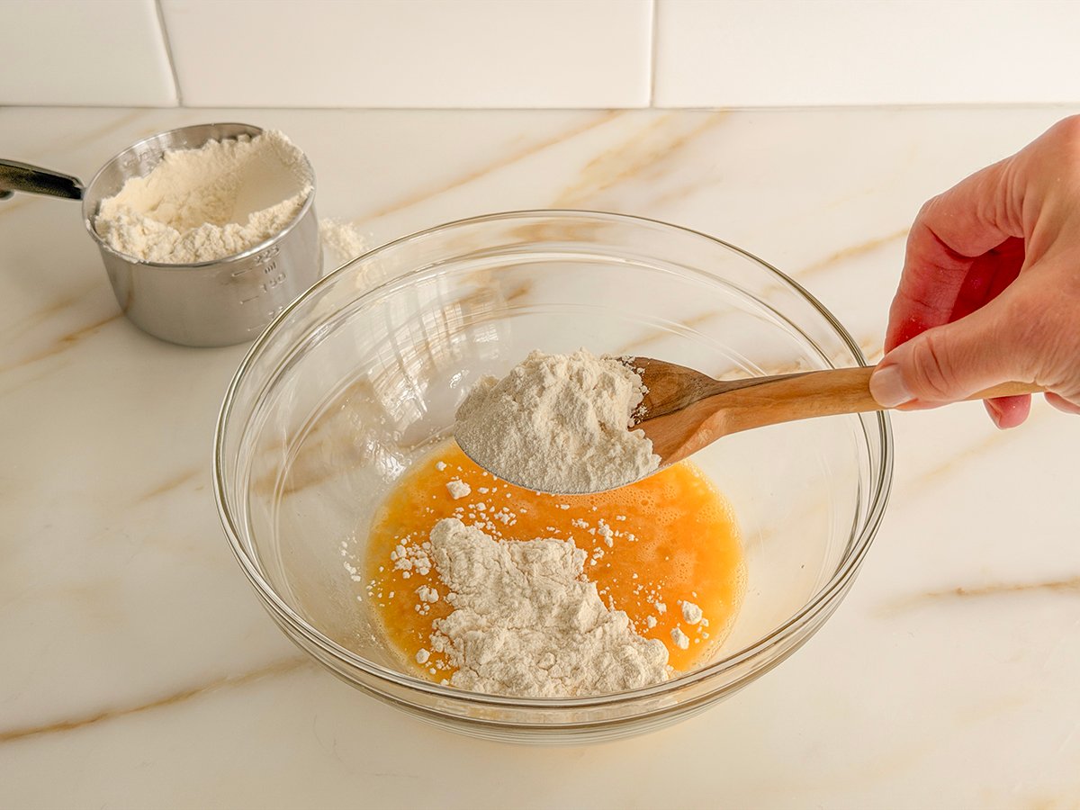 A wooden spoonful of flour over a bowl of whisked eggs