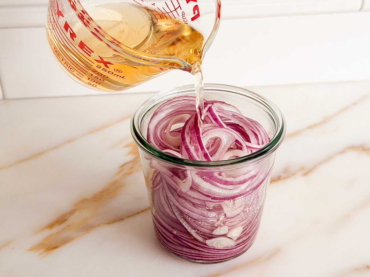 A liquid measure pouring amber liquid into red onions in a jar
