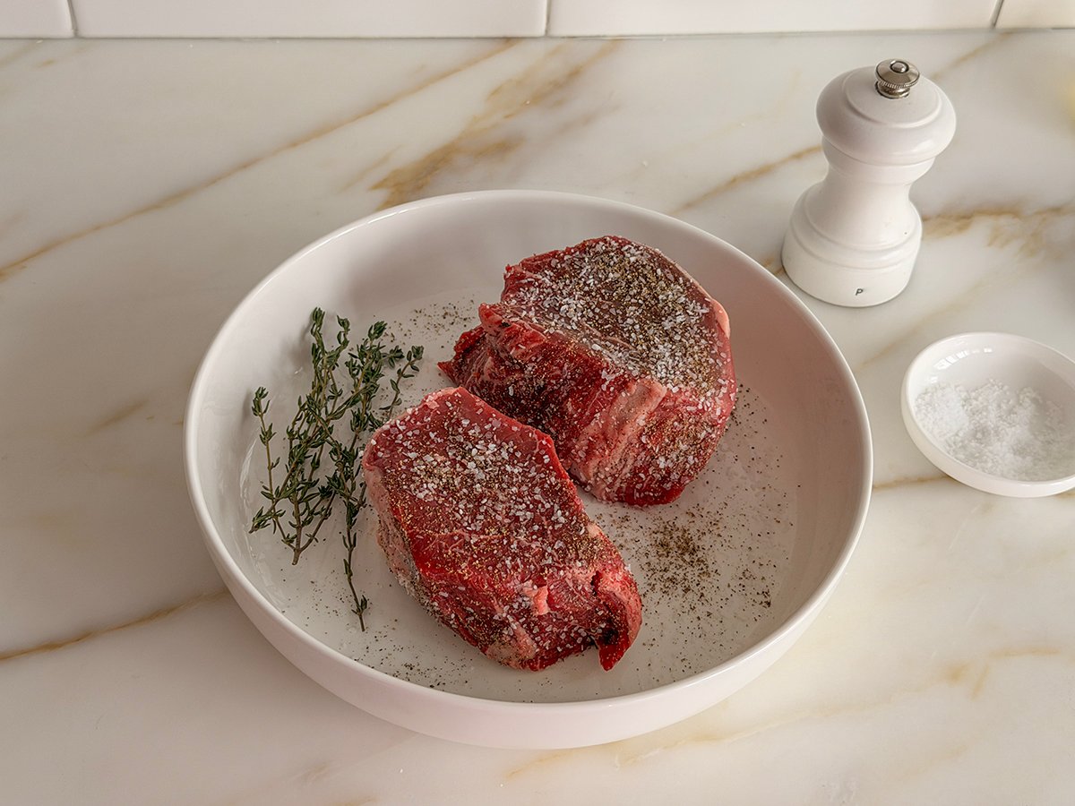 Seasoned filet mignon in a white dish with herbs on the side