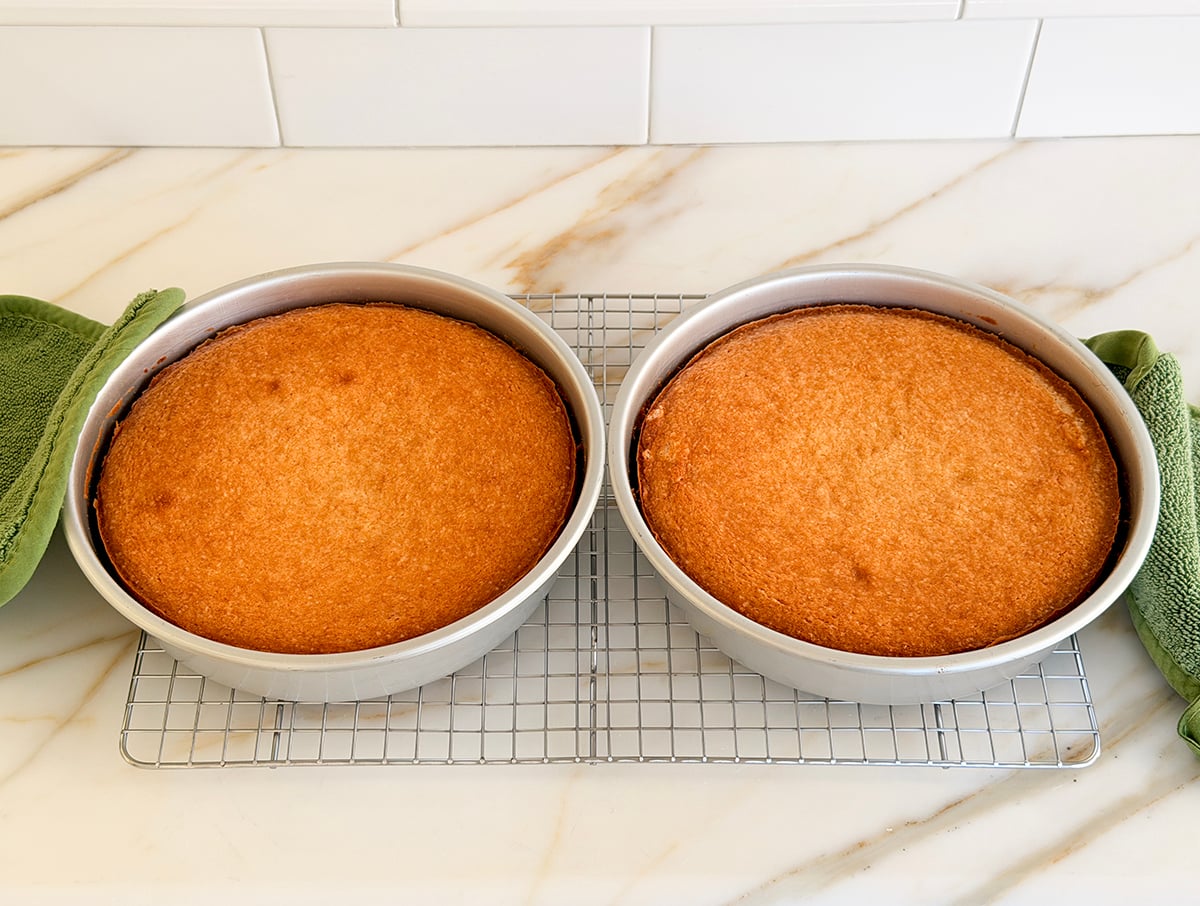 Two golden brown cakes in their round pans on a cooling rack with green hotpads