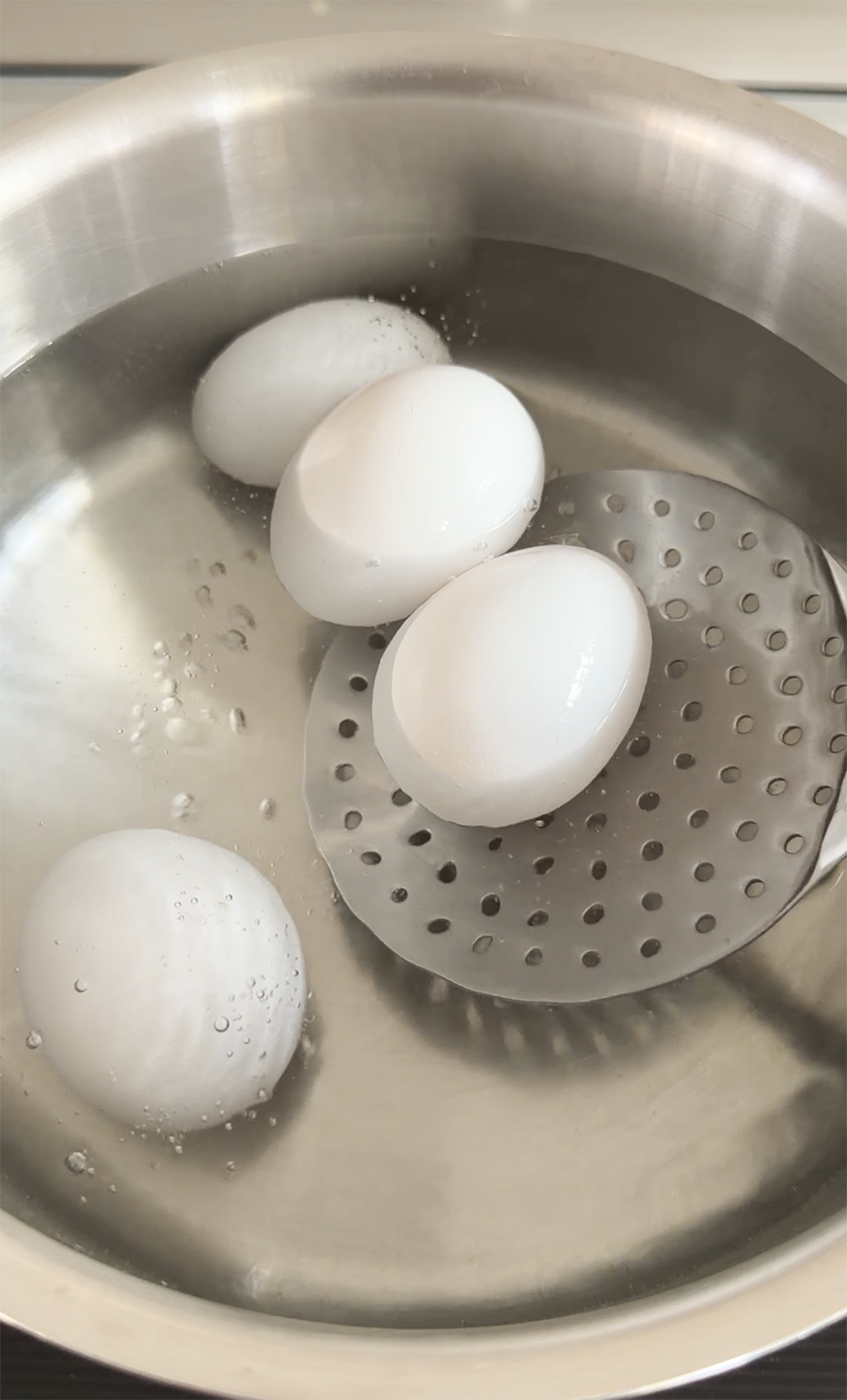 Eggs being added to water in a stainless steel pot with a slotted spoon.