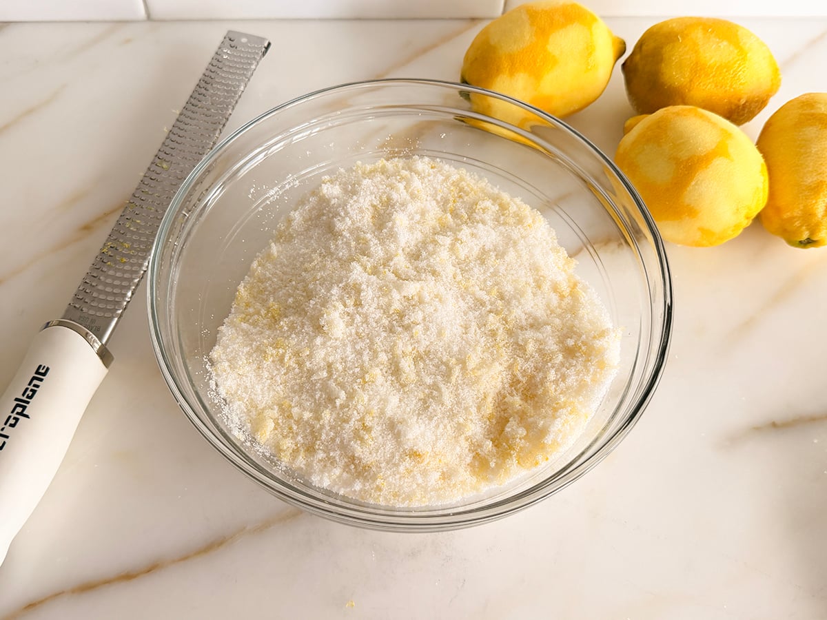 A glass bowl filled with damp sugar with lemons and a zester on the side