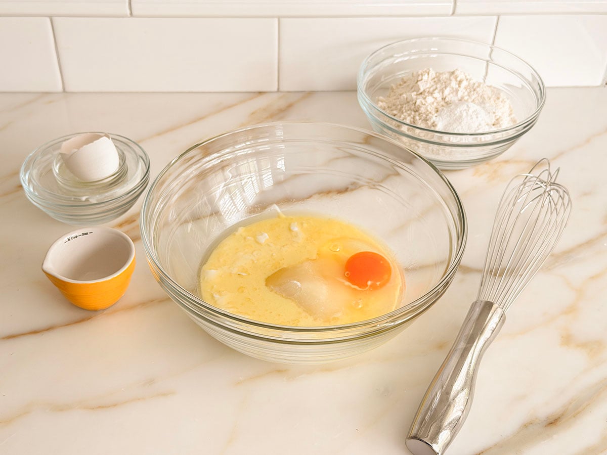 Glass bowls filled with an egg and other wet ingredients with glass bowl with flour on the side with a whisk