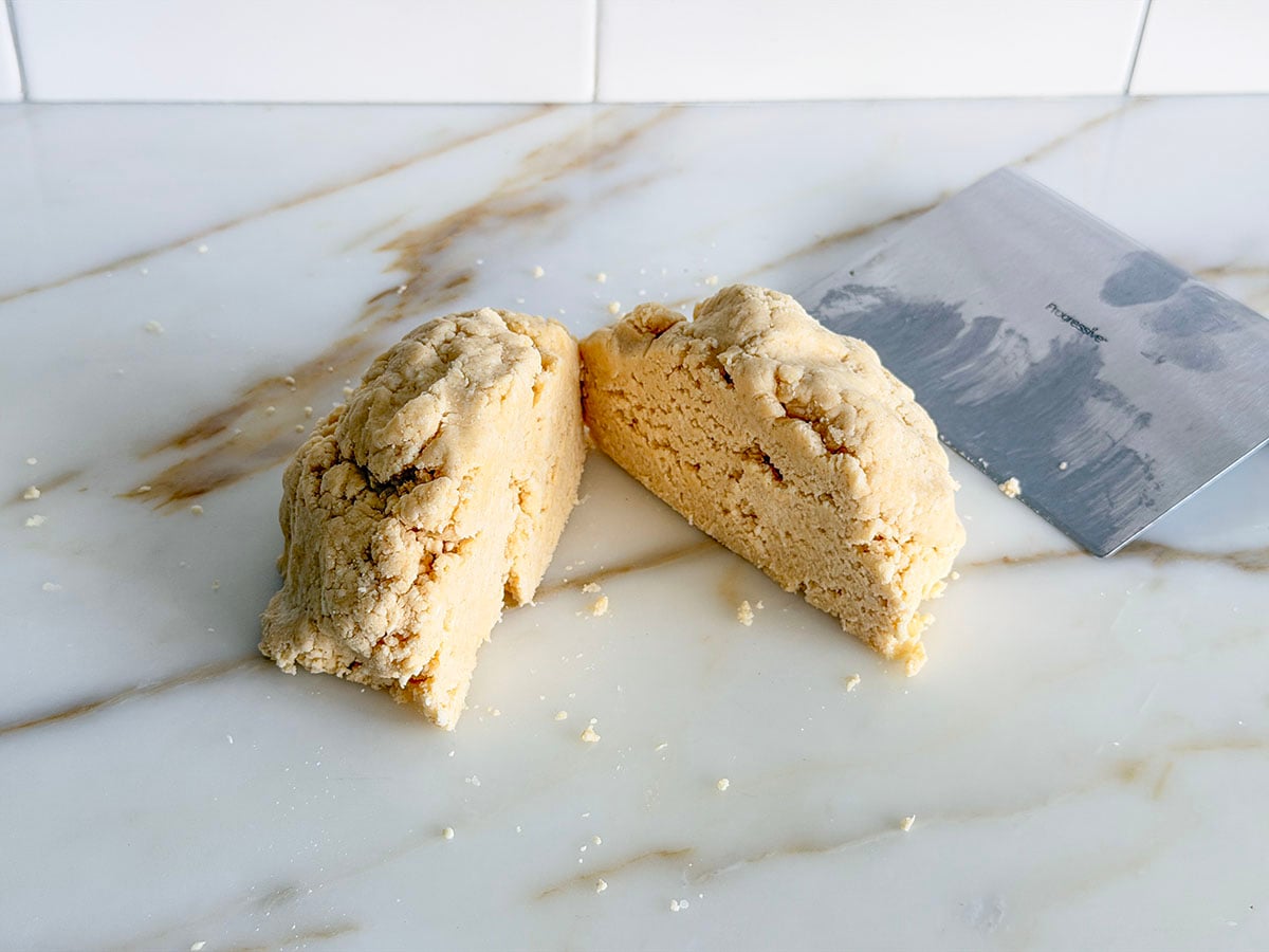 Pie dough ball cut in half on a marble countertop with bench scraper on the side