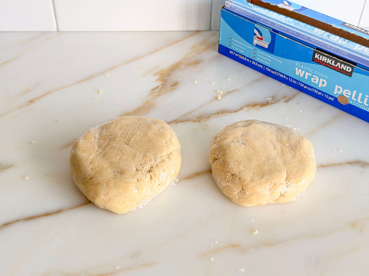 Two disks of pie dough wrapped in plastic on a marble countertop